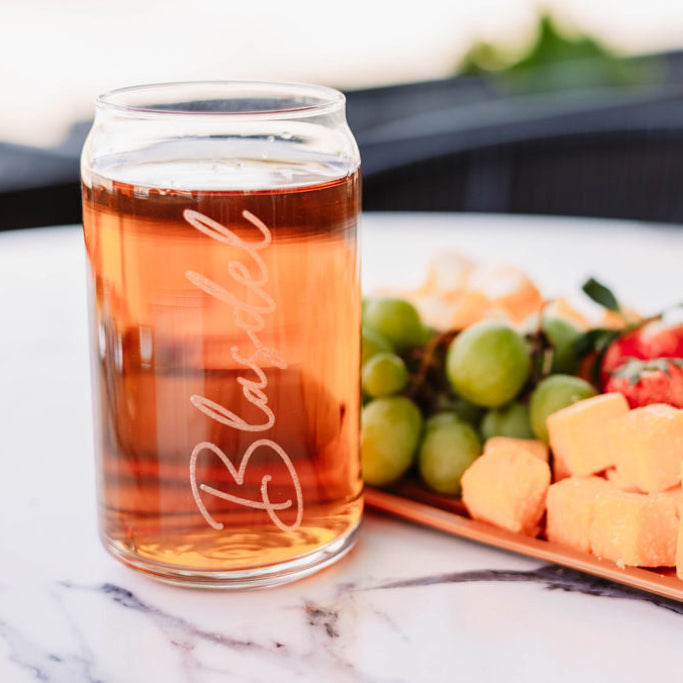 A glass with a engraved name on it, filled with a beverage, positioned next to a platter of fruits and cheese cubes.