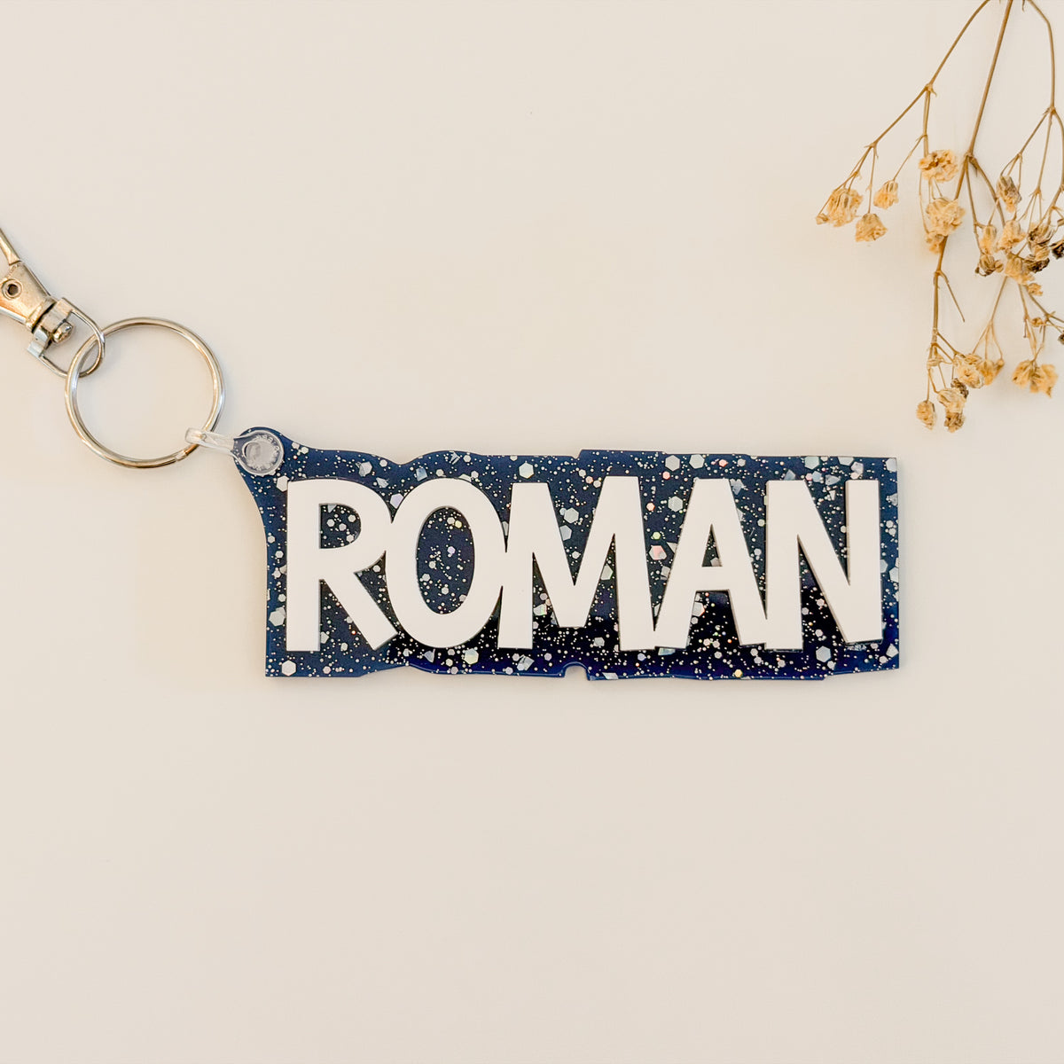 A rectangular keychain with the name 'ROMAN' in bold white letters on a dark navy blue background speckled with silver glitter, attached to a silver split keyring.