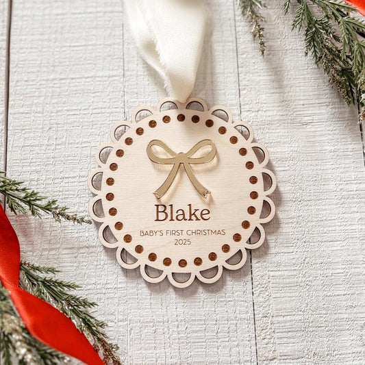 Decorative Christmas ornament with 'Blake' and 'Baby's First Christmas 2023' text on a white wooden surface with greenery and a red ribbon.