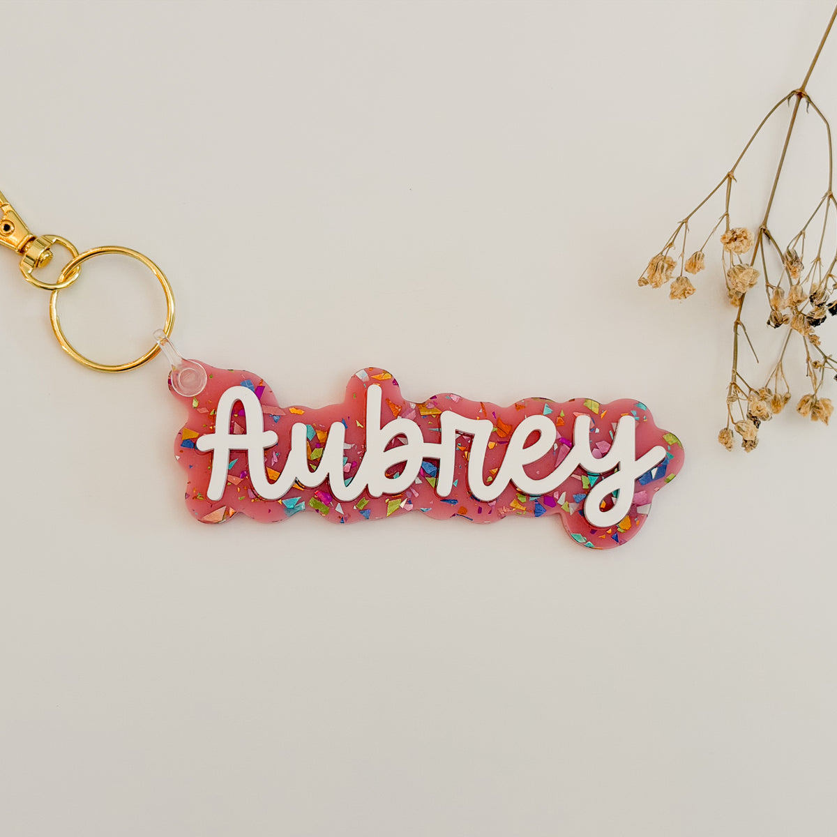 A pink acrylic name keychain with the name 'Aubrey' in white script, filled with multicolor glitter, attached to a gold key ring. 