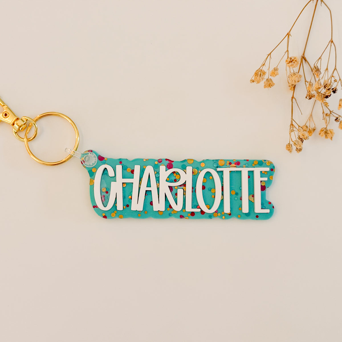 A personalized acrylic keychain with the name 'CHARLOTTE' in white letters on a turquoise background with multicolored confetti, attached to a gold key ring.