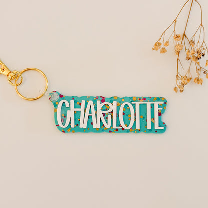 A personalized acrylic keychain with the name 'CHARLOTTE' in white letters on a turquoise background with multicolored confetti, attached to a gold key ring.