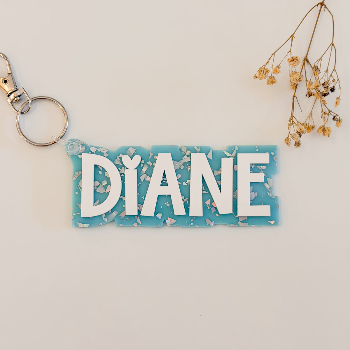 A personalized acrylic keychain with the name 'DIANE' in white letters on a light blue background with white shell-like flakes, attached to a silver split ring.