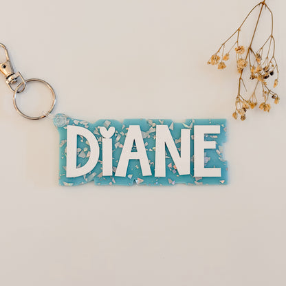 A personalized acrylic keychain with the name 'DIANE' in white letters on a light blue background with white shell-like flakes, attached to a silver split ring.