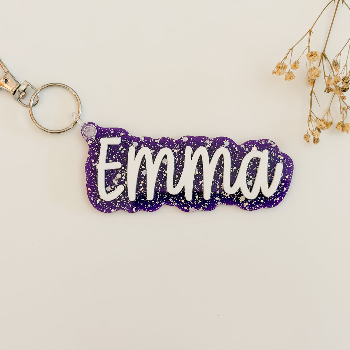 A custom name keychain featuring the name 'Emma' in white script on a purple glitter background, attached to a silver metal split ring and clasp.