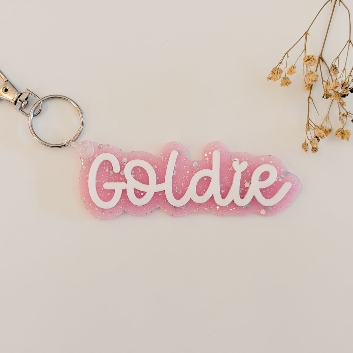 A pink glittery acrylic keychain with the name 'Goldie' in white cursive script, attached to a silver split ring and clasp.