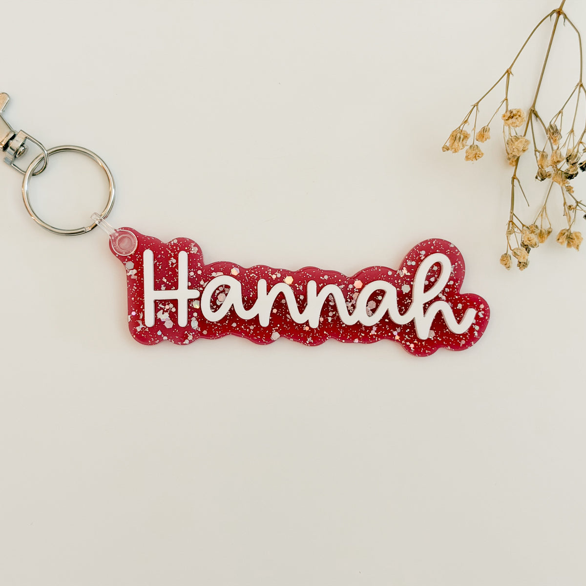 A red glittery acrylic keychain with the name 'Hannah' in white cursive script, attached to a silver split ring.