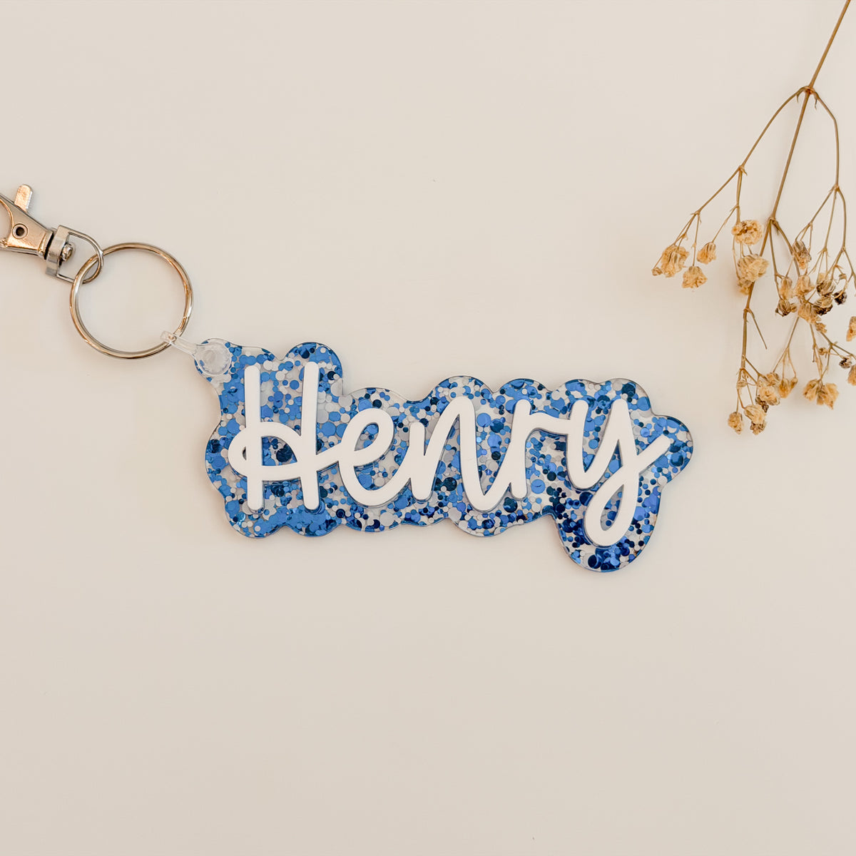 A custom name keychain featuring the name 'Henry' in white cursive script over a blue glittery background, attached to a silver split ring and clasp.