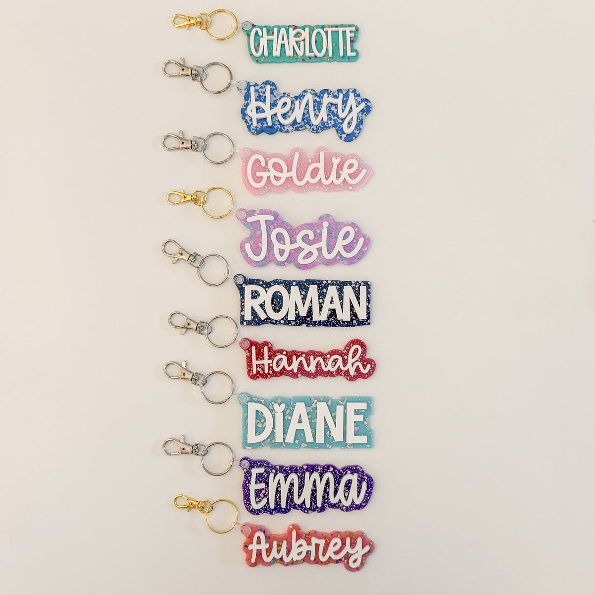 Nine personalized glitter name keychains arranged vertically on a white background, each with a gold split ring and clasp and names including Charlotte, Henry, Goldie, Josie, Roman, Hannah, Diane, Emma, and Aubrey.