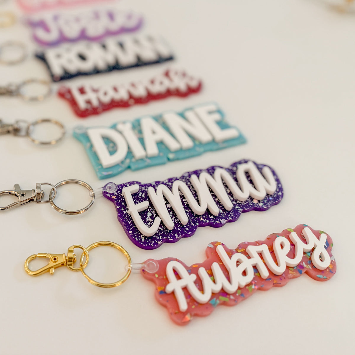 Five personalized name keychains  with silver keychains followed by a gold keychain and a pink glittery keychain spelling Aubrey, arranged diagonally on a light background.