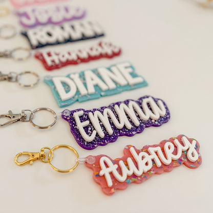 Five personalized name keychains  with silver keychains followed by a gold keychain and a pink glittery keychain spelling Aubrey, arranged diagonally on a light background.
