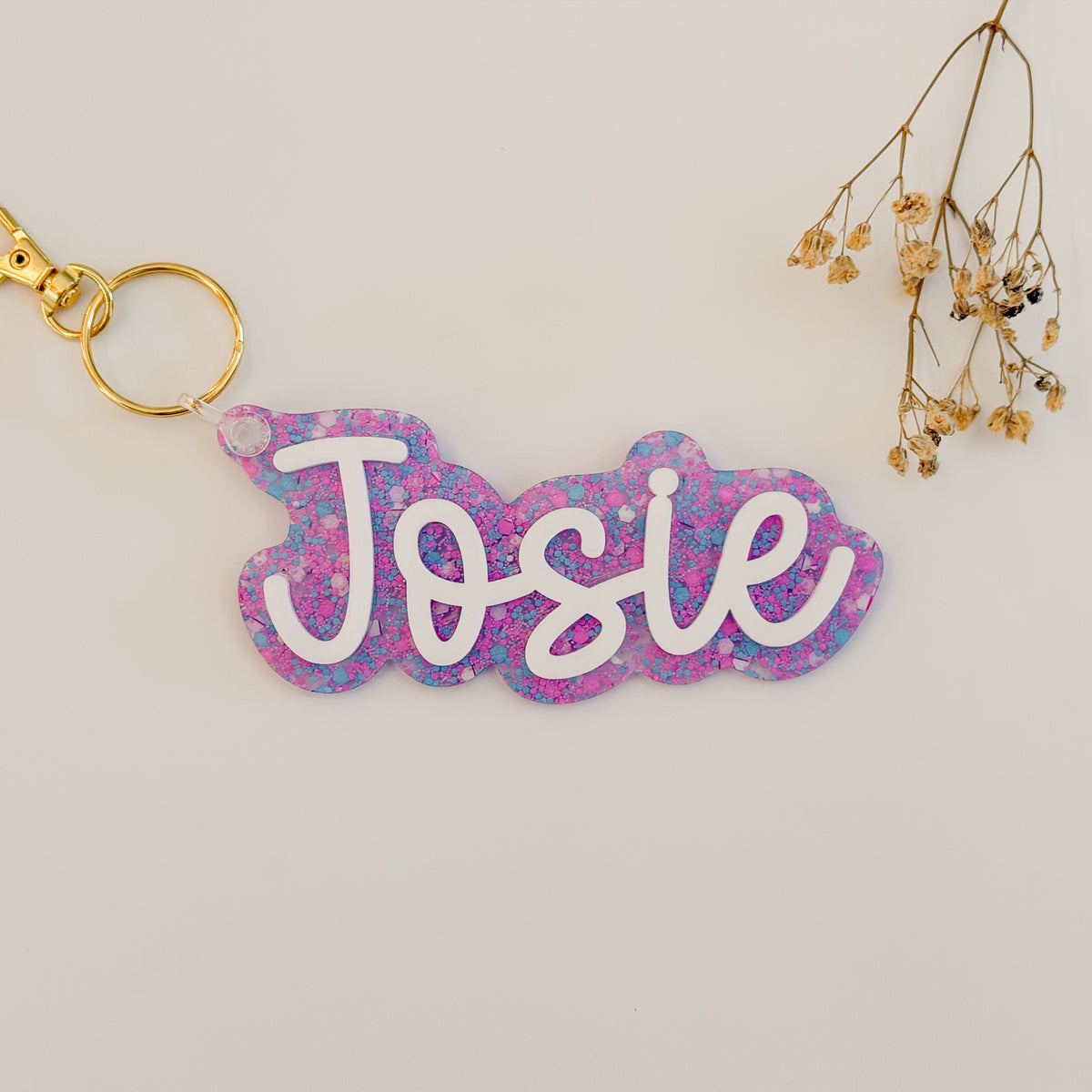 A personalized acrylic keychain with the name 'Josie' in white cursive lettering over a pink and blue glitter background, attached to a gold key ring.